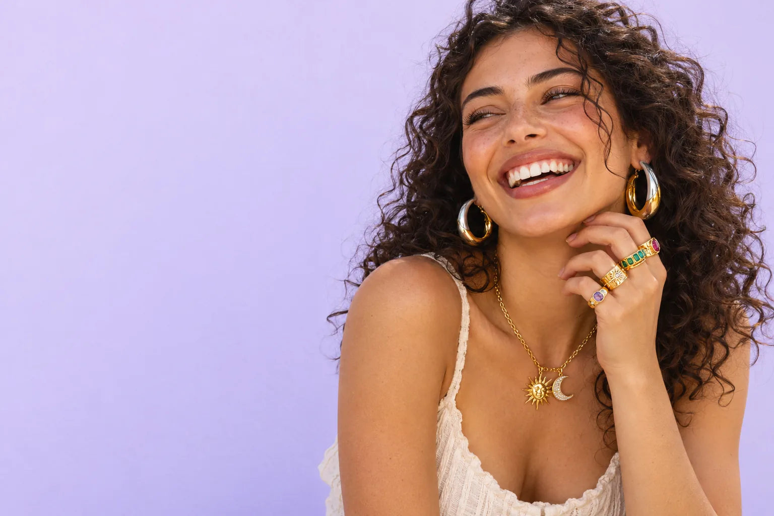 Woman laughing, wearing gold two-tone hoops, a celestial necklace and stacked colorful rings